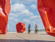 Zwei Radfahrer fahren zwischen großen orangefarbenen geometrischen Skulpturen auf einer Promenade in Ostende. Blauer Himmel mit weißen Wolken und Strand im Hintergrund.