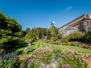 Kruidtuin Botanical Garden in Leuven with striped lawn, colorful flower beds, glass conservatory, and brick building under blue sky.