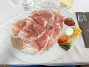 Weißer Teller mit dünn geschnittenem San Daniele Schinken, Mozzarella, getrockneter Tomate, Gurken, Kirschtomaten und Polenta. Weinglas sichtbar.