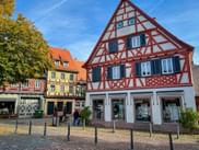 Traditionelles rot-weißes Fachwerkhaus in Ladenburg mit blauen Fensterläden auf einem Kopfsteinpflasterplatz. Bunte Gebäude im Hintergrund.