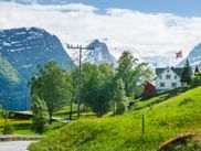 Weiße und rote Holzhäuser auf grünem Hang mit norwegischer Flagge. Schneebedeckte Berge und Gletscher unter bewölktem Himmel.