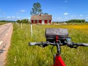 Rotes Fahrrad mit schwarzer Lenkertasche auf Radweg. Gelbes Wildblumenfeld und rotes schwedisches Häuschen mit weißen Fenstern im Hintergrund.