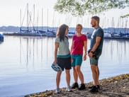 Three cyclists standing at the shore of Starnberger See in Bavaria, with a marina full of sailboats in the background. One person holds a helmet.
