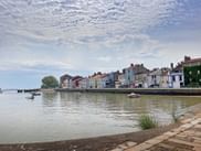 Row of colorful houses along the Loire River waterfront in Paimboeuf. Small boats float on the calm water under a cloudy sky.