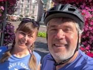 Selfie of Marcus Gloger in cycling helmet and a woman with braids in Bozen, framed by pink flowers with historic buildings in background.