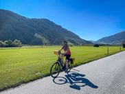 Female cyclist on paved path beside green meadow near Hollersbach, with forested mountains and blue sky in background.
