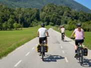 Three cyclists with panniers riding on a paved path near Kobarid, surrounded by green meadows and forested mountains under blue sky.