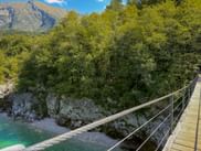 Wooden suspension bridge with rope railings crossing over turquoise Soča River surrounded by lush green forest and mountains near Kobarid.