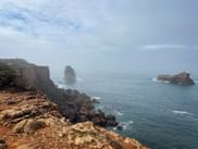 Felsige Küste bei Carrapateira mit dramatischen Klippen und zwei Felsformationen, die aus dem Ozean ragen, unter bewölktem Himmel.