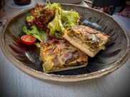 Zwei Stücke goldbrauner Lauchkuchen auf einem dunklen Keramikteller, serviert mit gemischtem Blattsalat und Cherrytomate in Bad Dürkheim.