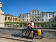 Radfahrerin mit Helm und gelben Taschen steht an Steinbalustrade und betrachtet die weiße barocke Villa Manin in Passariano unter blauem Himmel.