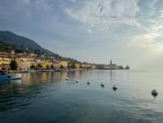 Waterfront view of Salò on Lake Garda with colorful buildings, church tower, mountains, and buoys floating in calm water under blue sky.