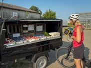 Radfahrerin mit Helm und Rucksack steht an einem Obststand am Straßenrand mit Beeren in Behältern. Eine Scheune und ein Gewächshaus sind sichtbar.