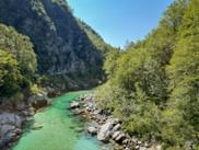 Turquoise Soča River flowing through a rocky gorge near Kobarid, surrounded by green trees and steep mountain slopes under blue sky.