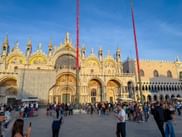 St. Mark's Basilica in Venice with golden mosaics and Gothic spires. Crowds gather in the square with two tall red flagpoles visible.