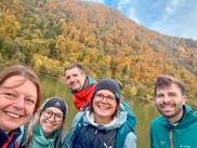 Fünf lächelnde Personen in Outdoorjacken machen ein Selfie an der Donau mit herbstlich gefärbten Hügeln im Hintergrund bei einem Betriebsausflug.