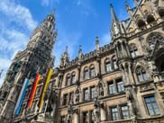 Munich New Town Hall with ornate Gothic facade, tall central tower, and colorful flags against blue sky.