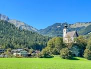 White church with dark roof and tower in Golling surrounded by green meadows, traditional houses, and forested alpine mountains under clear blue sky.
