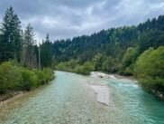 Turquoise Isar River with gravel banks flowing through forested valley near Mittenwald. Dense coniferous forests cover the hillsides under cloudy sky.