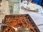 Metallschale mit Cargols a la llauna, gegrillten Schnecken in würziger Sauce, auf weißer Tischdecke mit Weingläsern und Tellern in Katalonien.