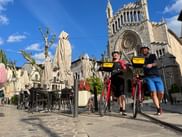 Two cyclists with red bikes stand in front of Sóller Cathedral in Mallorca. The Gothic church facade towers behind them on a sunny plaza.