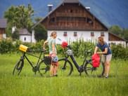 Zwei Radfahrer mit Kind machen Pause auf einer Wiese vor einem traditionellen österreichischen Bauernhaus im Salzkammergut mit Tourenrädern.