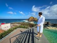 Person in white bathrobe standing by pool in Porto Moniz, Madeira, overlooking Atlantic Ocean with rocky island and coastal facilities under blue sky.