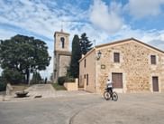 Radfahrer mit Fahrrad vor einer historischen Steinkirche mit Glockenturm in Katalonien unter blauem Himmel mit weißen Wolken.