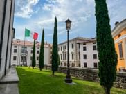 Courtyard in Udine with green lawn, tall cypress trees, Italian flag, and historic buildings under a blue sky with white clouds.