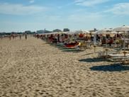Breiter Sandstrand in Caorle mit Reihen weißer Sonnenschirme und Liegestühlen. Strandbesucher entspannen sich auf dem Sand unter blauem Himmel mit Gebäuden im Hintergrund.