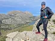Smiling hiker with backpack standing on rocky outcrop overlooking Cuber reservoir in Mallorca's Tramuntana mountains under blue sky.