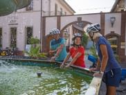 Drei Radfahrer mit Helmen erfrischen sich an einem Brunnen im Zentrum von Deidesheim, mit historischen Gebäuden und Torbogen im Hintergrund.