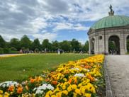 Diana Temple pavilion with green copper dome in Munich Hofgarten. Colorful flower beds with yellow and orange blooms line the path to the temple.