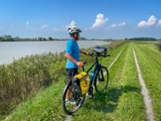 Radfahrer mit blauem Shirt und Helm steht mit Tourenrad auf Grasweg neben Wasserweg. Gelbe Packtaschen am Rad befestigt.