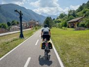 Cyclist with yellow panniers riding on paved bike path through Chiusaforte, with green mountains and village buildings in the background.
