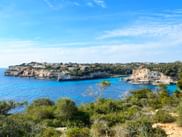 Bucht mit türkisfarbenem Wasser und Kalksteinklippen nahe Es Pontàs, Mallorca. Grüne Vegetation im Vordergrund, weiße Gebäude auf Klippen unter blauem Himmel.
