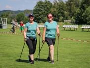 Two women in matching turquoise shirts with Nordic walking poles on a grass field at Grabenseelauf 2026. White benches and trees in background.