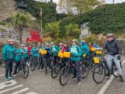Große Gruppe von Radfahrern in türkisfarbenen Jacken mit Helmen und Fahrrädern mit gelben Taschen vor Steinmauer mit Palmen.