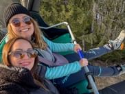 Zwei lächelnde Frauen mit Sonnenbrillen und Winterjacken fahren am Wintersporttag 2026 mit einem Sessellift über einen verschneiten Wald.