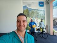 Smiling man in turquoise jacket taking selfie at Eurobike office in Bozen with cycling poster showing mountain landscape in background.