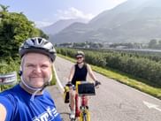 Two cyclists with helmets on a bike path with mountains in the background. Man in blue shirt taking selfie, woman on red bike behind him.