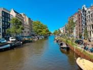 Amsterdamer Gracht gesäumt von traditionellen niederländischen Häusern und Booten an beiden Seiten unter blauem Himmel mit grünen Bäumen.