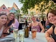 Vier Frauen machen ein Selfie an einem Tisch eines Straßencafés mit Getränken und Weinflaschen in Bad Dürkheim. Bunte historische Gebäude im Hintergrund.