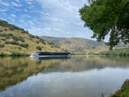 Modern river cruise ship on calm Douro River near Foz do Sabor, surrounded by hills with sparse vegetation under blue sky.