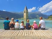 Seven people sitting on a wooden pier viewing the famous bell tower rising from Lake Reschen in South Tyrol, with mountains in the background.