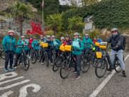 Große Gruppe von Radfahrern mit Helmen und gelben Packtaschen posiert mit ihren Rädern auf einer Straße. Österreichische Flagge und Denkmal im Hintergrund.