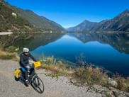 Cyclist with yellow panniers on path beside Lake Idro. Mountains reflect in calm blue water under clear sky.