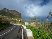 Road sign marking Porto da Cruz on a winding coastal road in Madeira, with mountains, vegetation, and dramatic cloudy sky in the background.