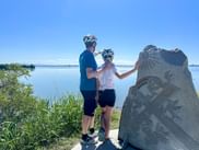 Two cyclists with helmets standing beside a stone monument with sun carving, overlooking the calm Adriatic Sea in Grado under clear blue sky.