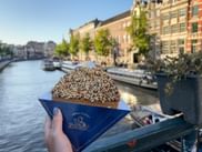 Hand hält eine Stroopwafel in blauer Verpackung entlang des Rokin-Kanals in Amsterdam mit historischen Gebäuden, Booten und Bäumen im Hintergrund.